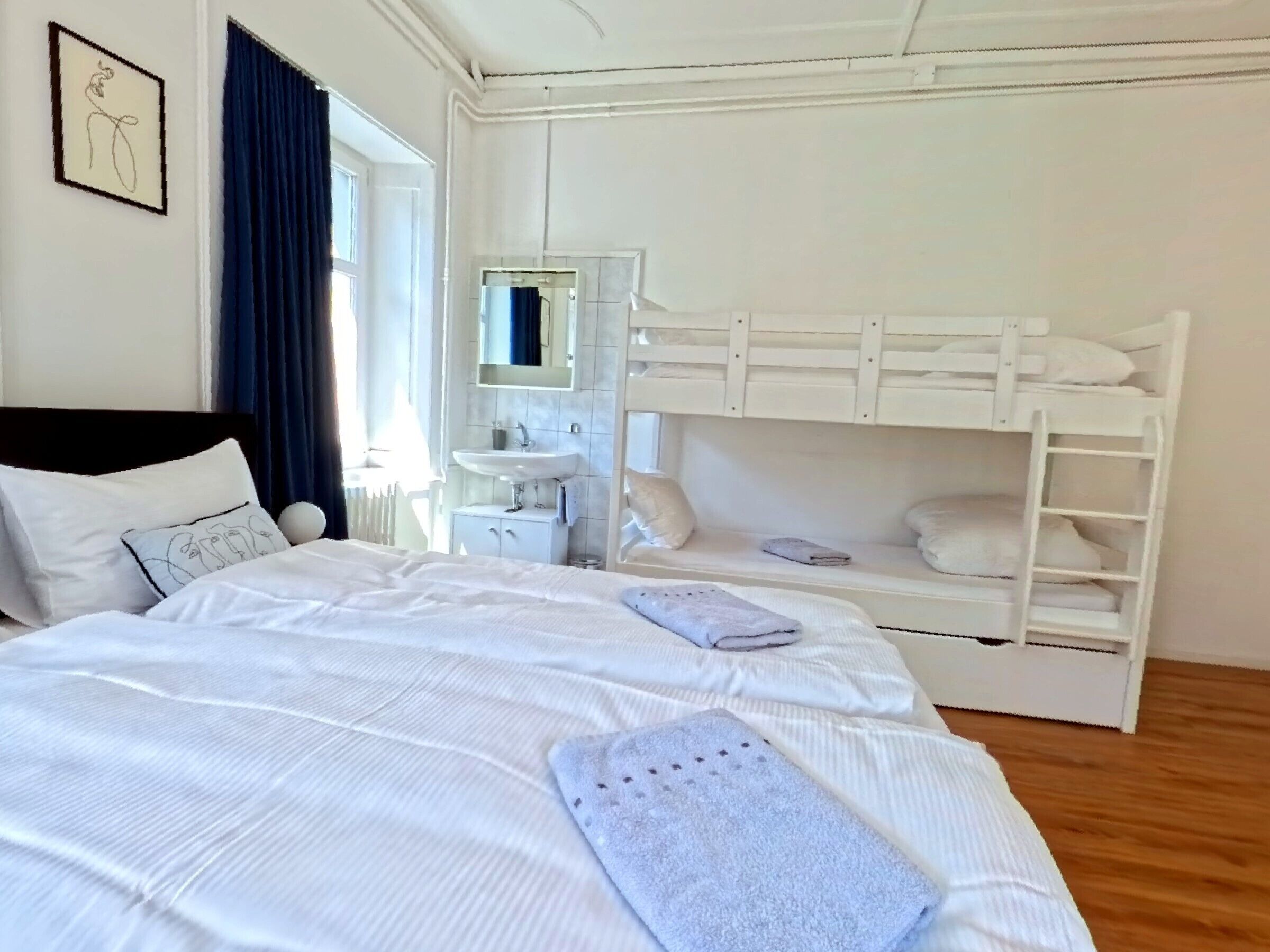 Una habitación de hotel con una cama, un tocador blanco, un espejo, una cama nido y una ventana con cortinas.