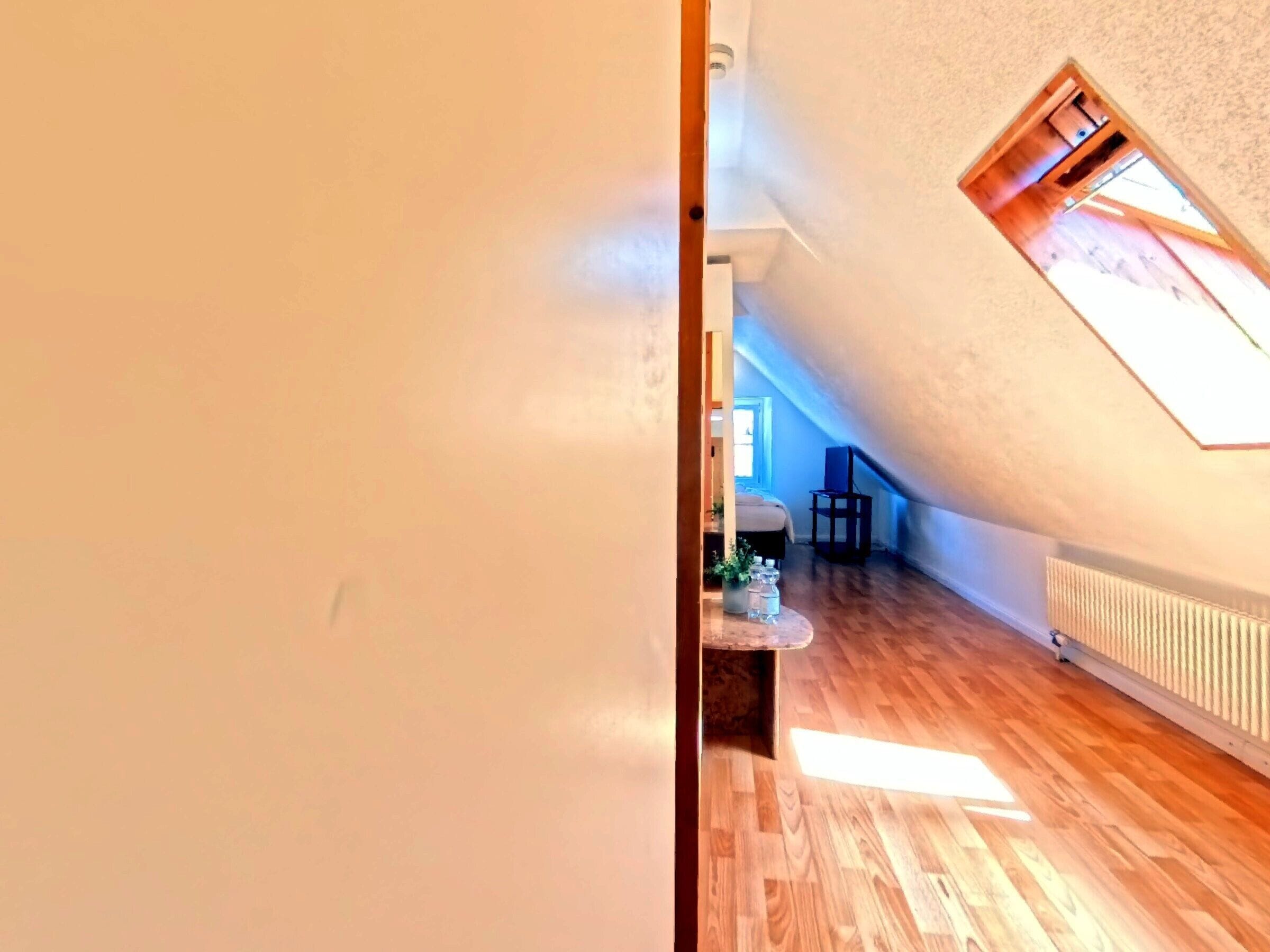 Ein schmaler Flur mit Holzboden führt zu einem Raum mit schrägem Dach und kleinem Fenster.