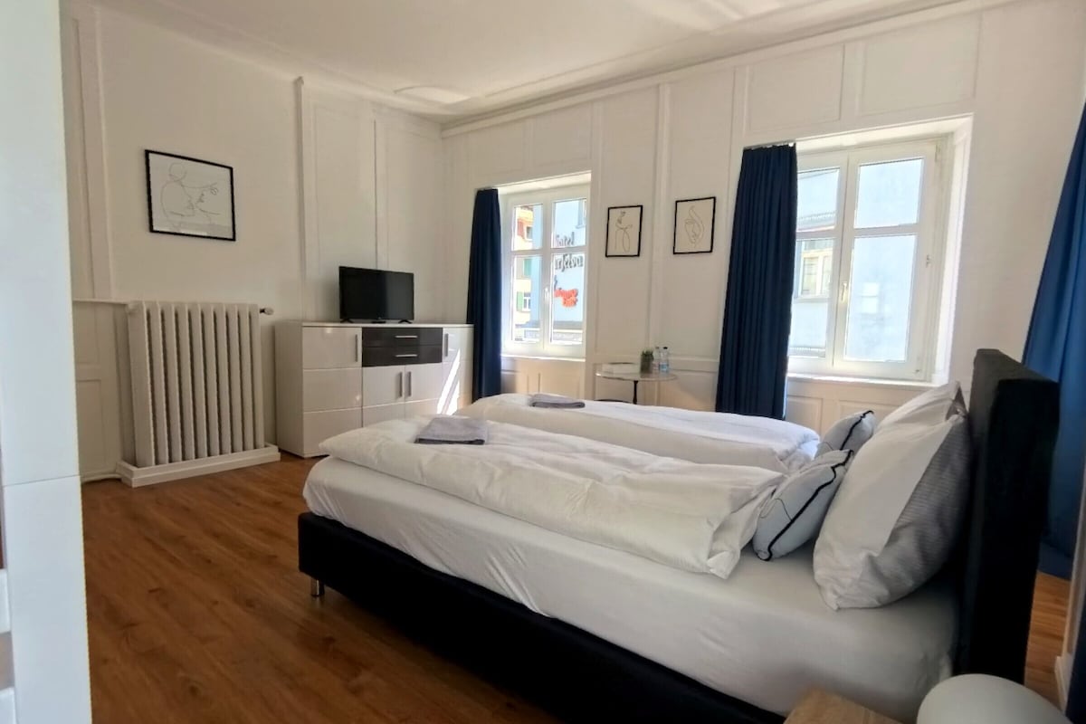 A hotel room with a double bed, a bedside table, a dresser, a radiator, and a window with curtains.