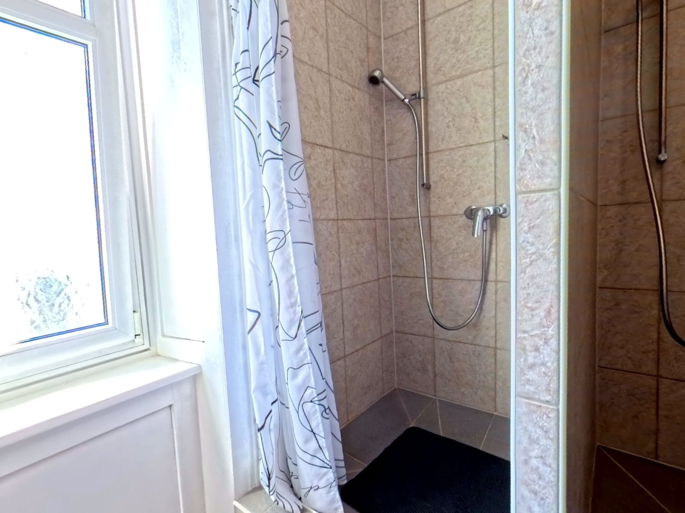 Una ducha con cabezal de ducha de mano y grifo empotrado en la pared, una ventana con marco blanco, y una puerta que da acceso a la zona de la ducha.
