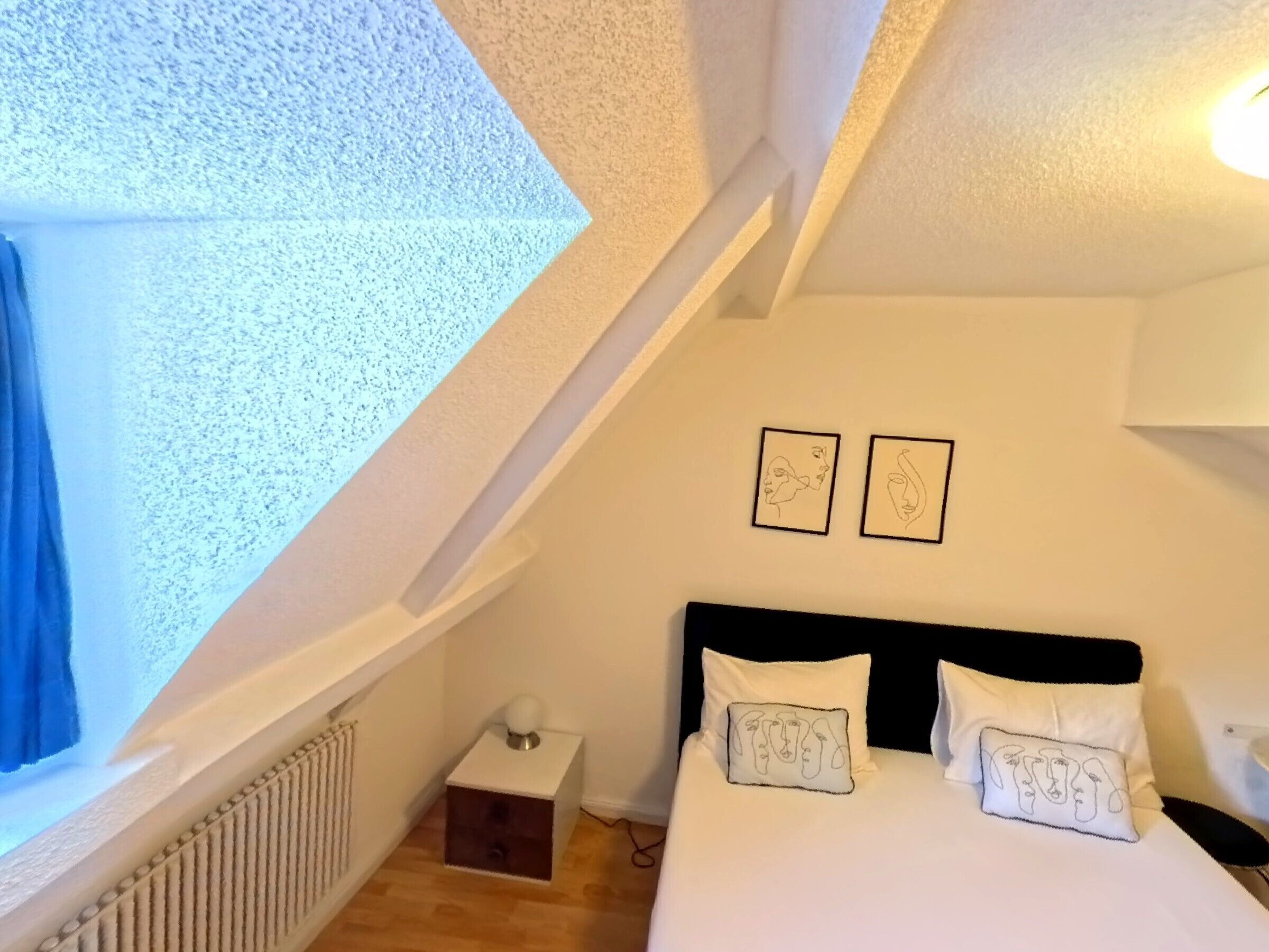 Un dormitorio con techo inclinado, una cama con ropa de cama blanca, dos cuadros en la pared y una mesita de noche con lámpara.