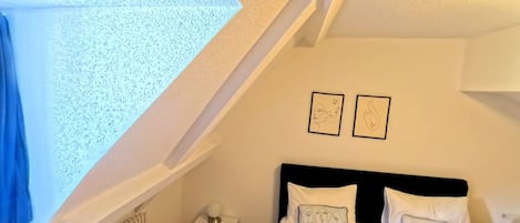 A bedroom with a sloped ceiling, a bed with white bedding, two wall-mounted pictures, and a small bedside table with a lamp.