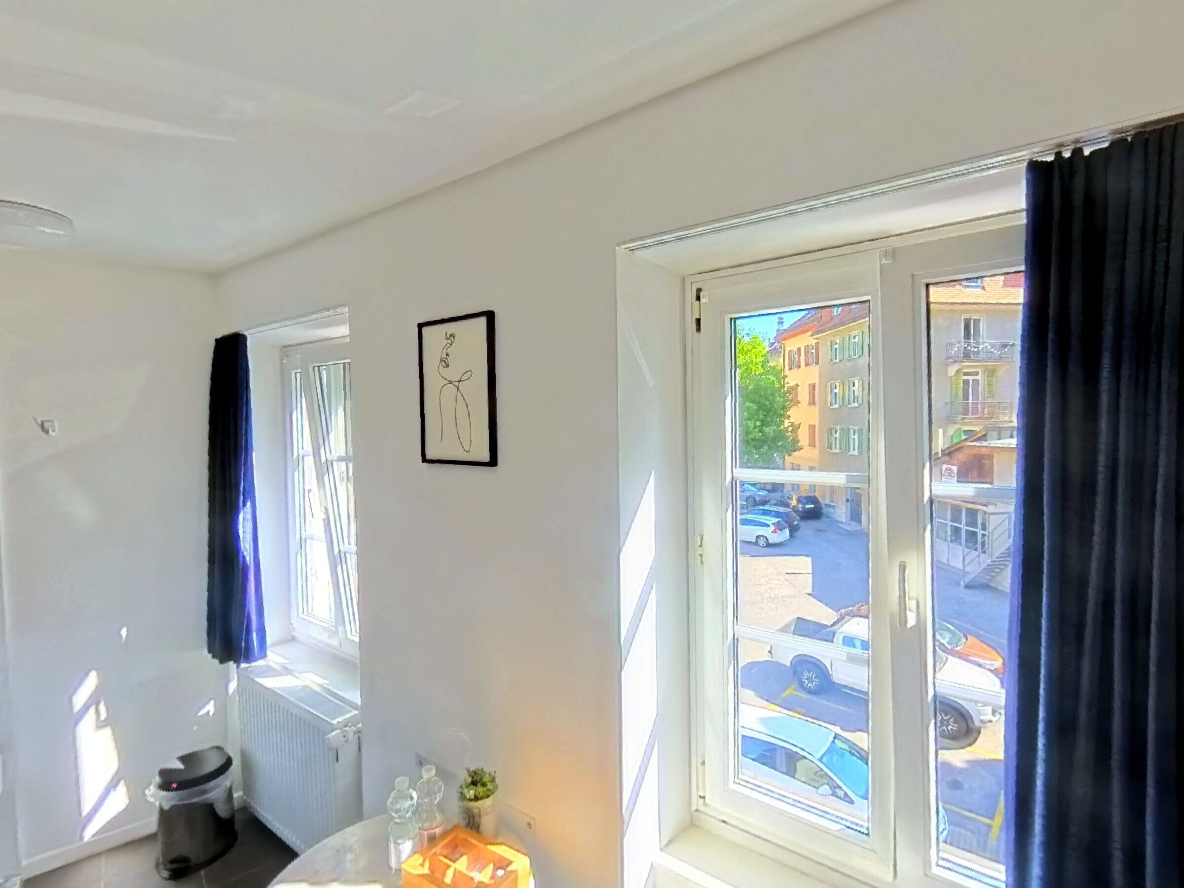 Una habitación con ventana, un radiador, un cubo de basura y un cuadro en la pared.