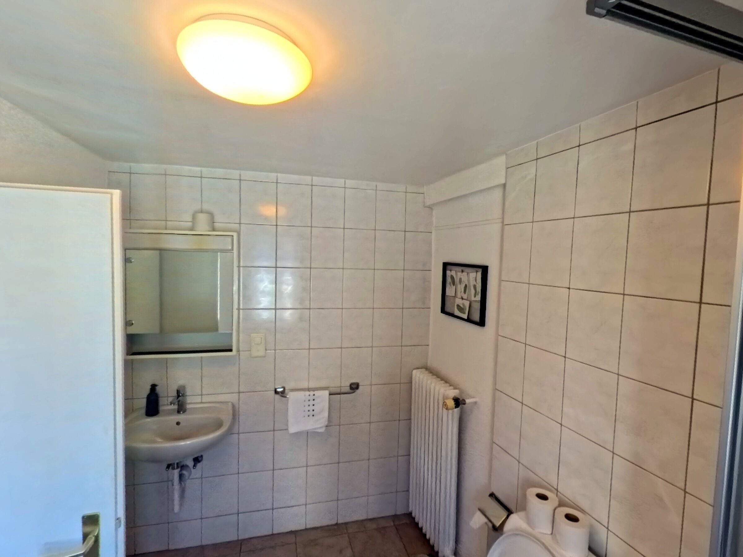 Ein Badezimmer mit weißen Fliesen, einem Waschbecken, einem Spiegel, einem Handtuchhalter und einer Heizung.
