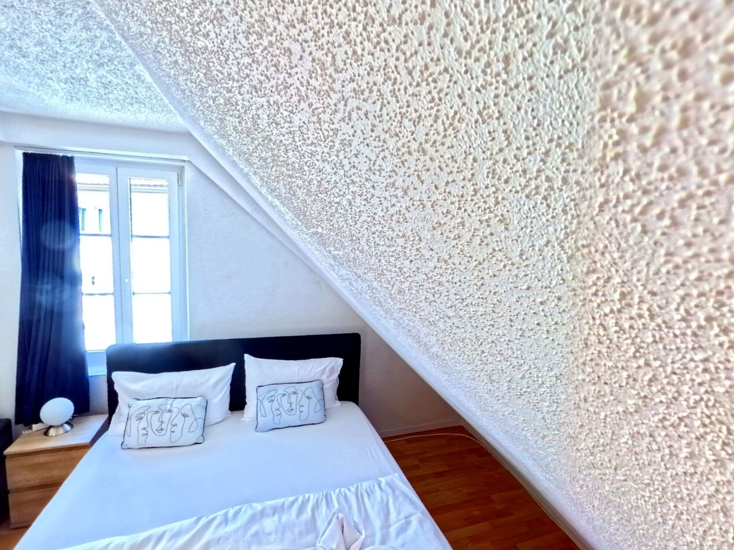 Un dormitorio con cama, dos almohadas, una mesita de noche y una ventana con cortinas.