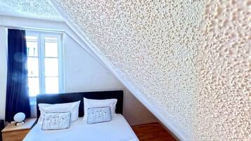 Un dormitorio con cama, dos almohadas, una mesita de noche y una ventana con cortinas.