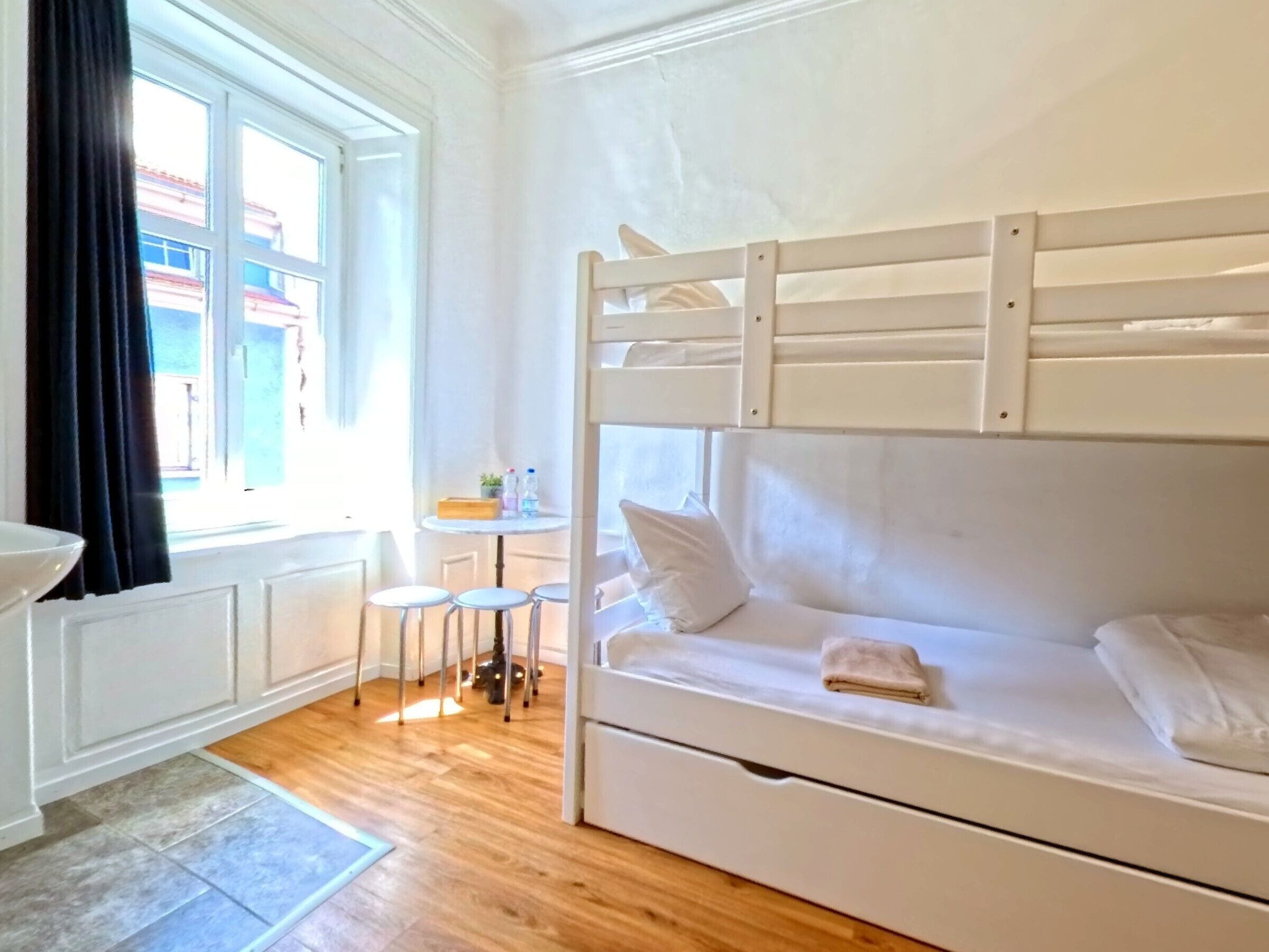 Una cama litera con una pequeña mesa y taburetes, una ventana con cortinas y vistas a edificios.