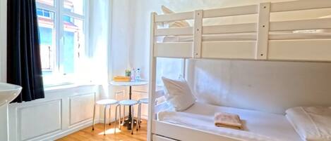 A bunk bed with a small table and stools, a window with curtains, and a view of buildings outside.