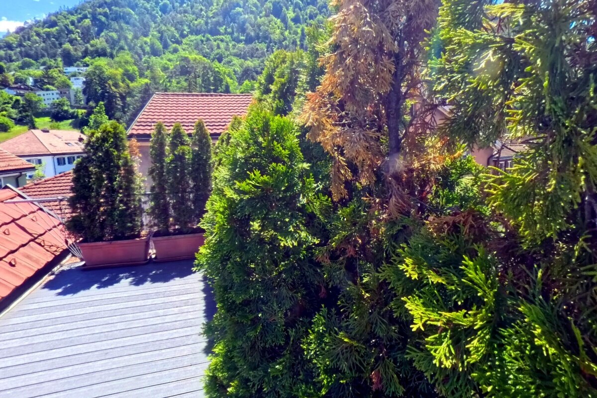 A residential area with a house featuring a red-tiled roof, surrounded by dense greenery and a hill in the background.