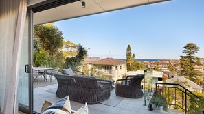 A balcony with wicker furniture, a glass door, and a view of a coastal town. - Salt Ridge North Bondi (North Bondi)