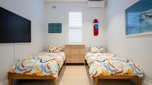 Ein Schlafzimmer mit zwei Betten, einer Kommode, einem an der Wand montierten Fernseher, einem Bild und einem Fenster mit Jalousien.