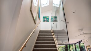 A staircase with glass railings leading to a second floor, with artwork on the walls and a ceiling fan. - Alora House, North Bondi (North Bondi)