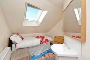 Un dormitorio con tragaluz, una cama con ropa de cama floral, una mesita de noche, una mesita de noche de madera, un lavamanos blanco y un espejo.
