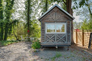 Une petite cabane en bois avec une fenêtre, entourée d’arbres et bordée d’un chemin de gravier.