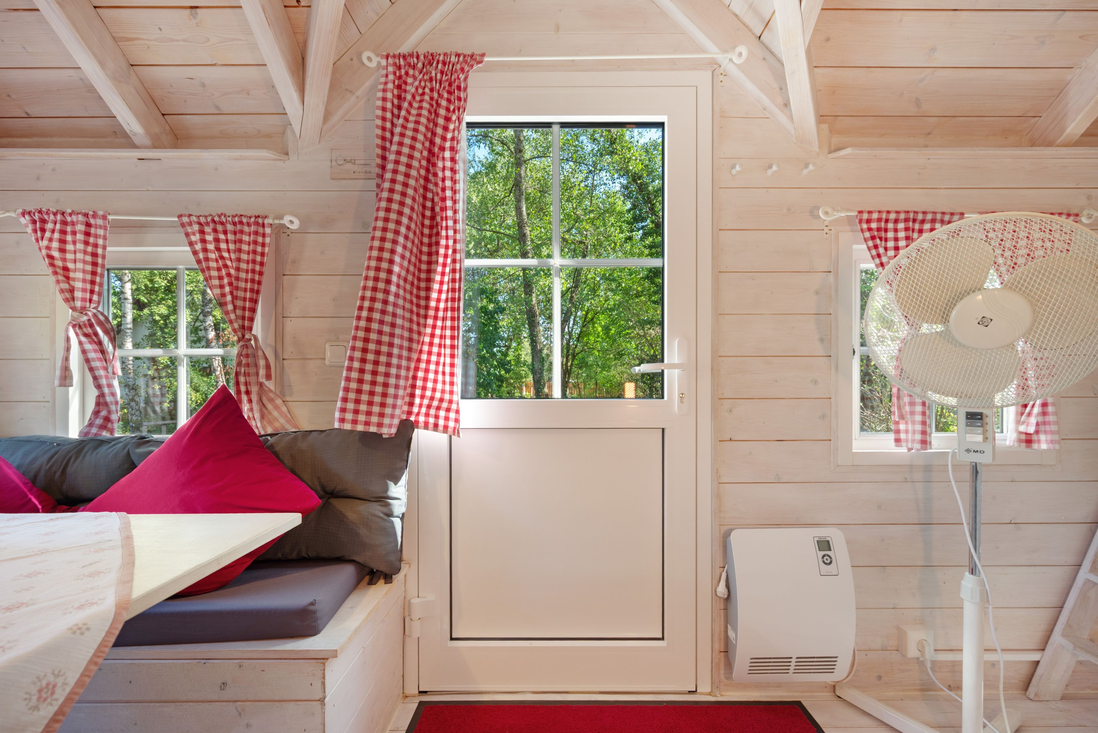A cozy room with a bed, a window with checkered curtains, a standing fan, and a heater.