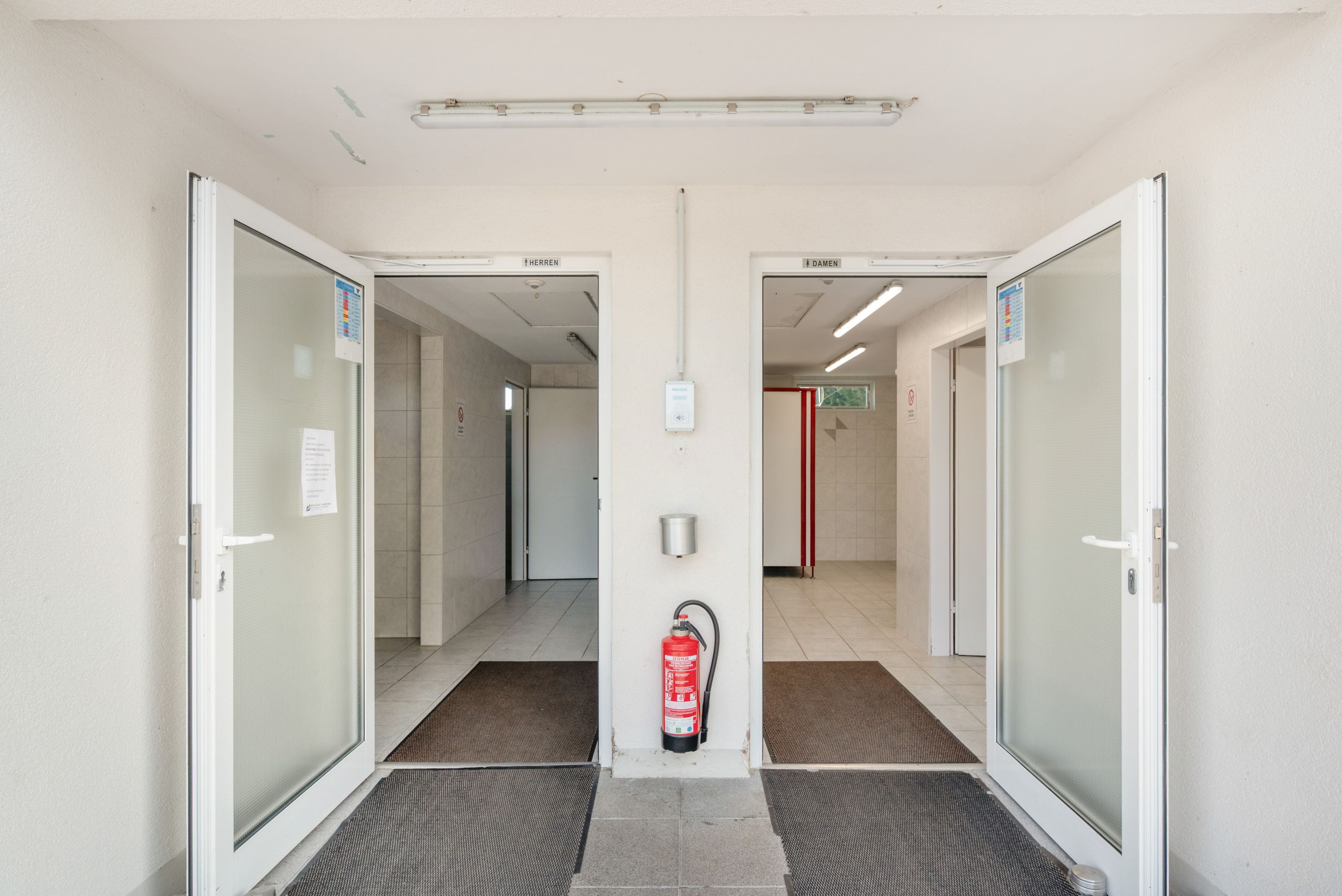 L’entrée d’un hôtel, avec deux portes portant les inscriptions « Herren » et « Damen », qui donnent sur un couloir carrelé.