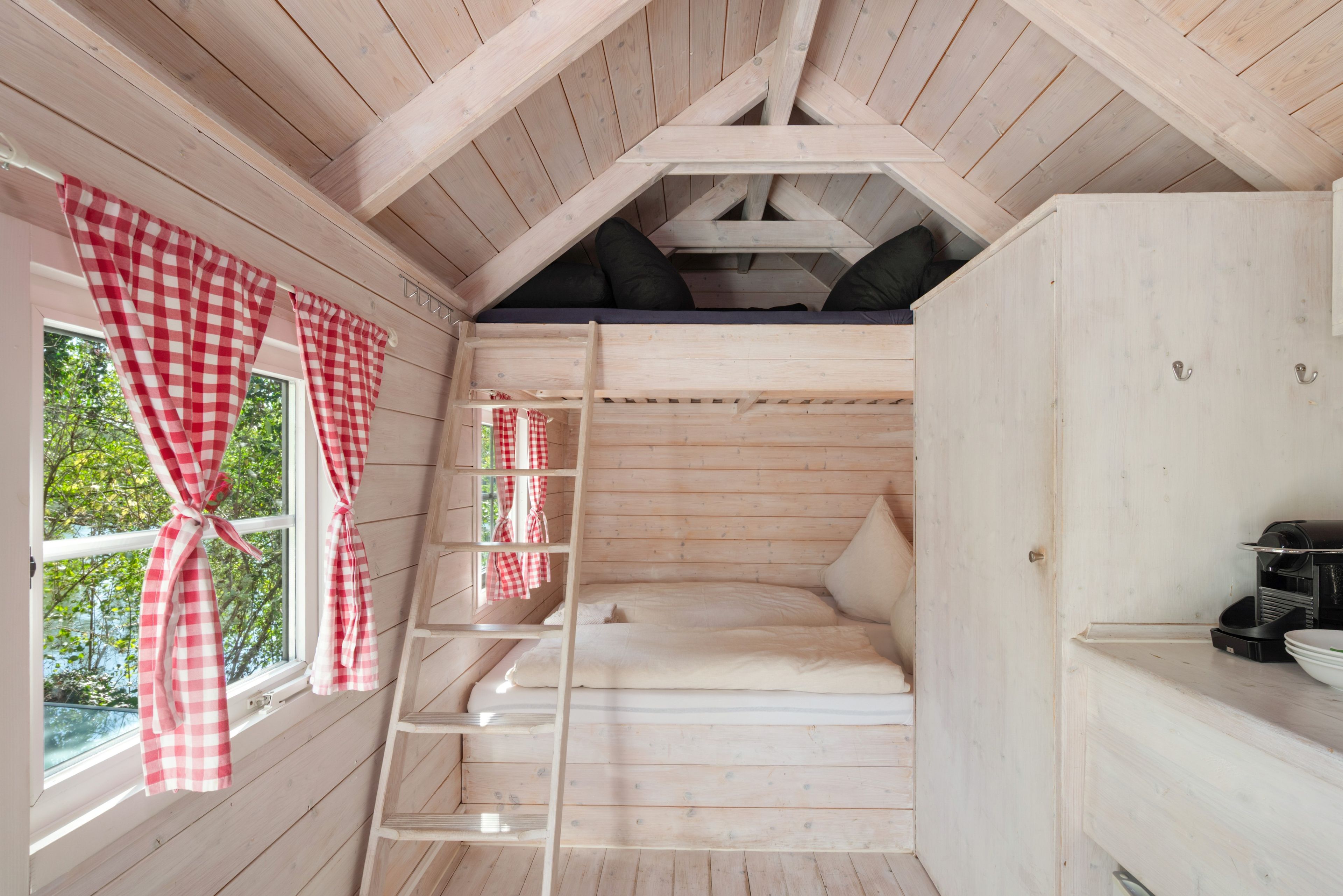 Eine Holzhütte mit Schlafkoje, einer Sprosse und einem Fenster mit karierten Vorhängen.