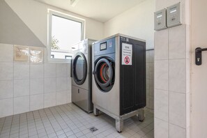 Une buanderie équipée de deux machines à laver et d’un sèche-linge, avec une fenêtre et un panneau indiquant qu’il est interdit d’y entrer avec des chiens.