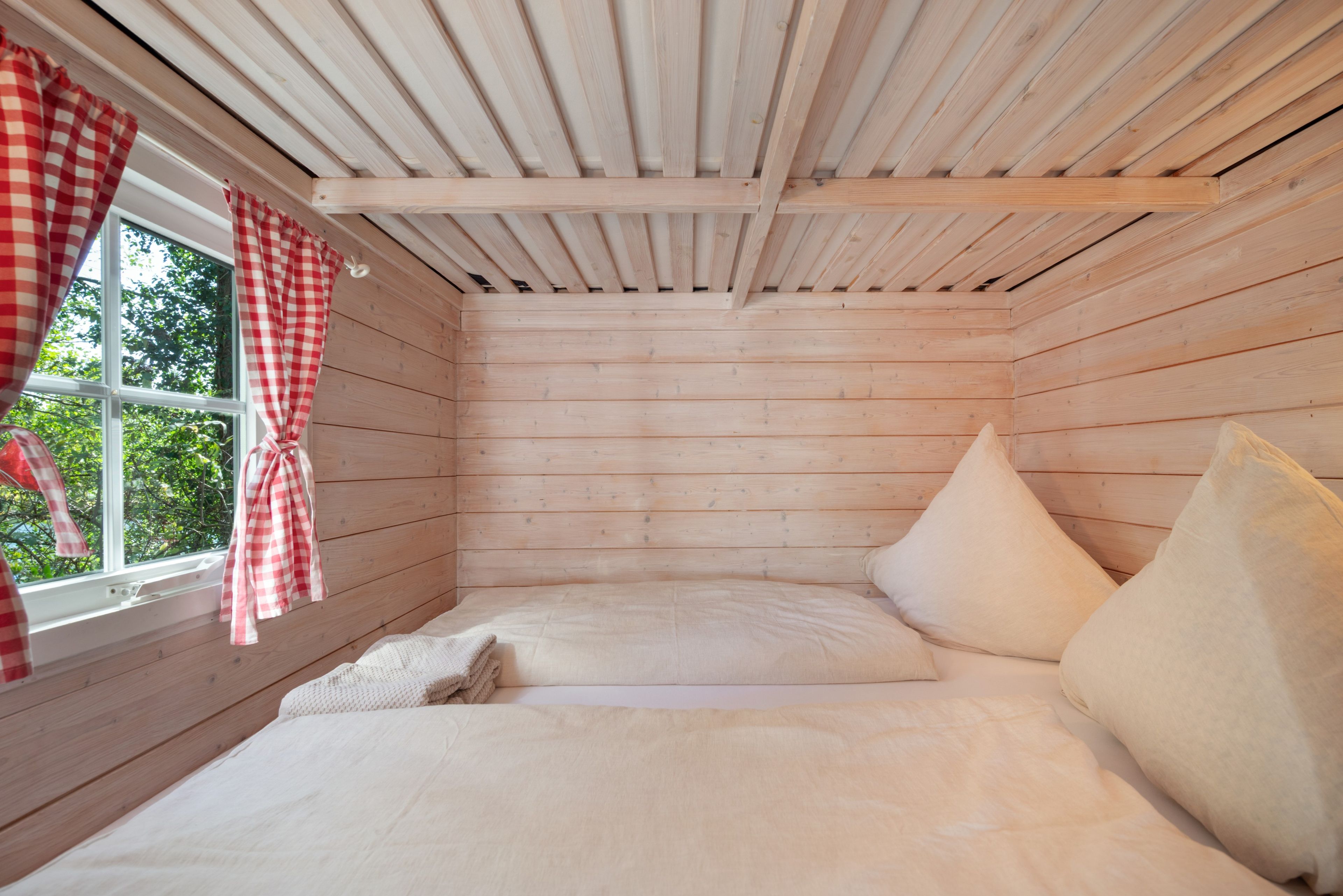 Ein Zimmer mit Holzwänden, einem Bett mit Kissen und einem Fenster mit Karomustervorhängen.