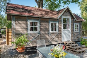 A rustic wooden cabin with a sloped roof, two windows with white frames, a door, and a small porch area with a metal chair and a table.