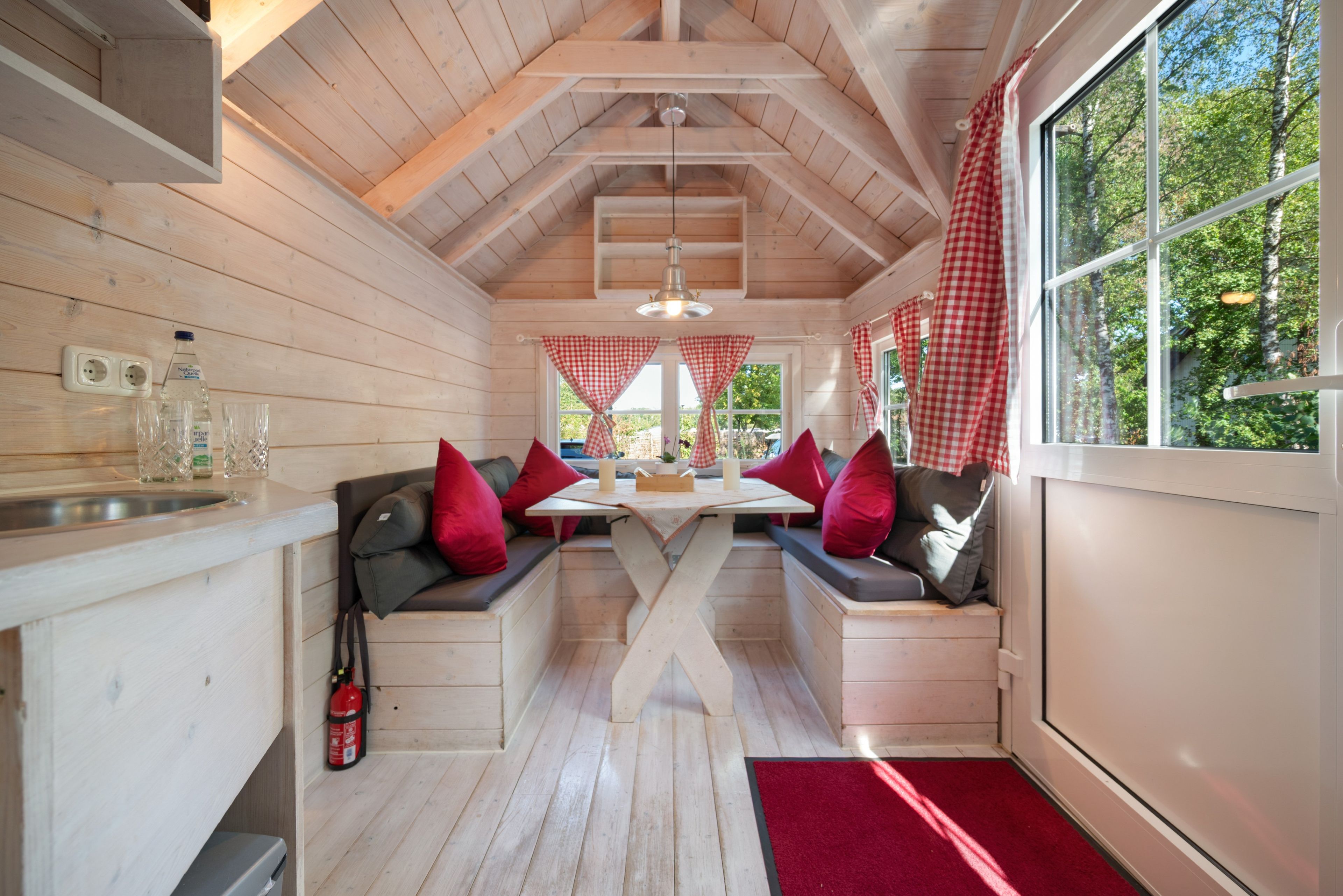 Ein Holzblockhaus-Innenraum mit Esstisch, Bänken und einem Fenster mit roten und weißen Karomustert-Vorhängen.