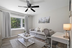 A modern living room with a ceiling fan, a sofa, a coffee table, a desk, and a lamp. - Pocono Oasis | Hot Tub • Game Room • Fenced Yard (Long Pond)