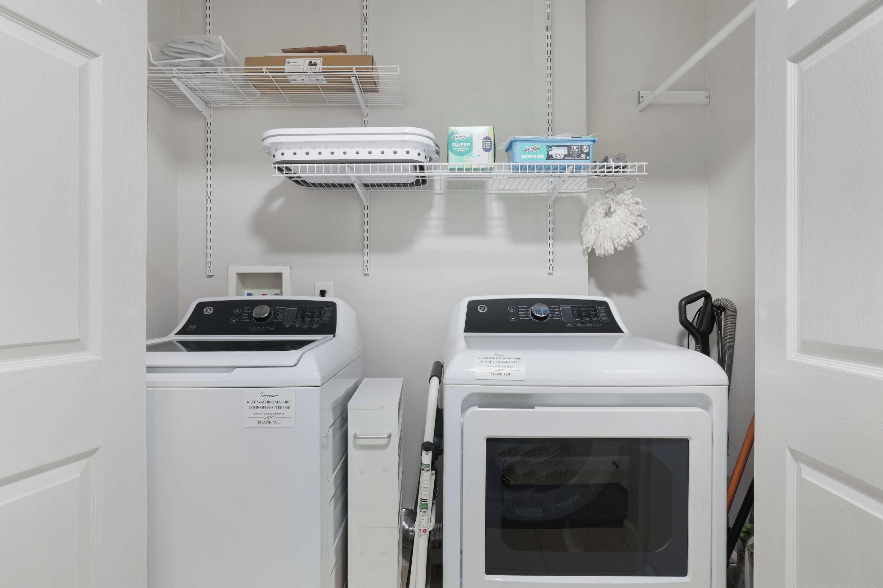 A laundry room with two front-loading washing machines, a dryer, and a shelf with cleaning supplies.