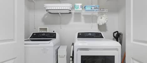 A laundry room with two front-loading washing machines, a dryer, and a shelf with cleaning supplies.