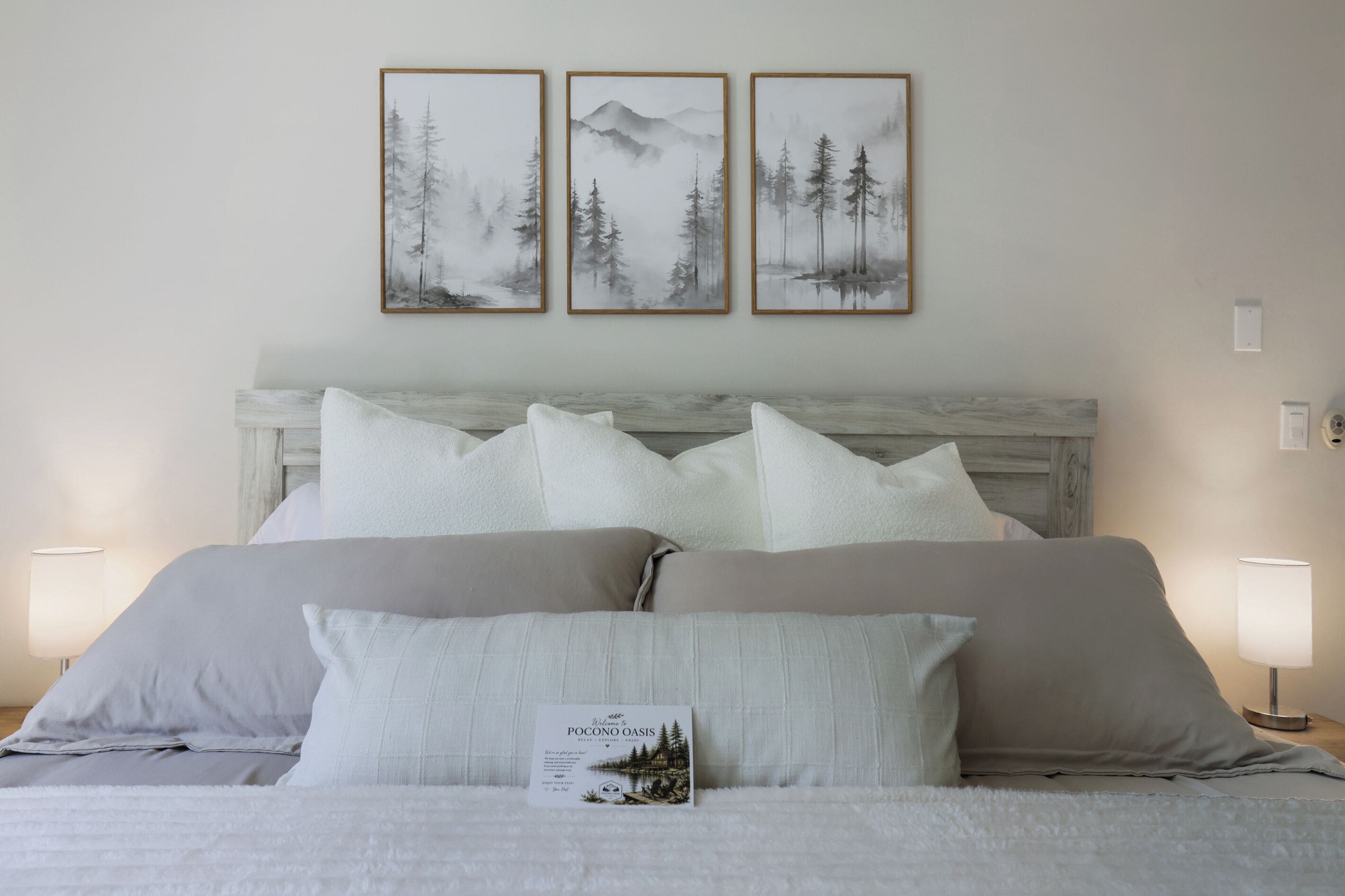 A bed with white linens and pillows, a wooden headboard, and three framed landscape paintings on the wall above.