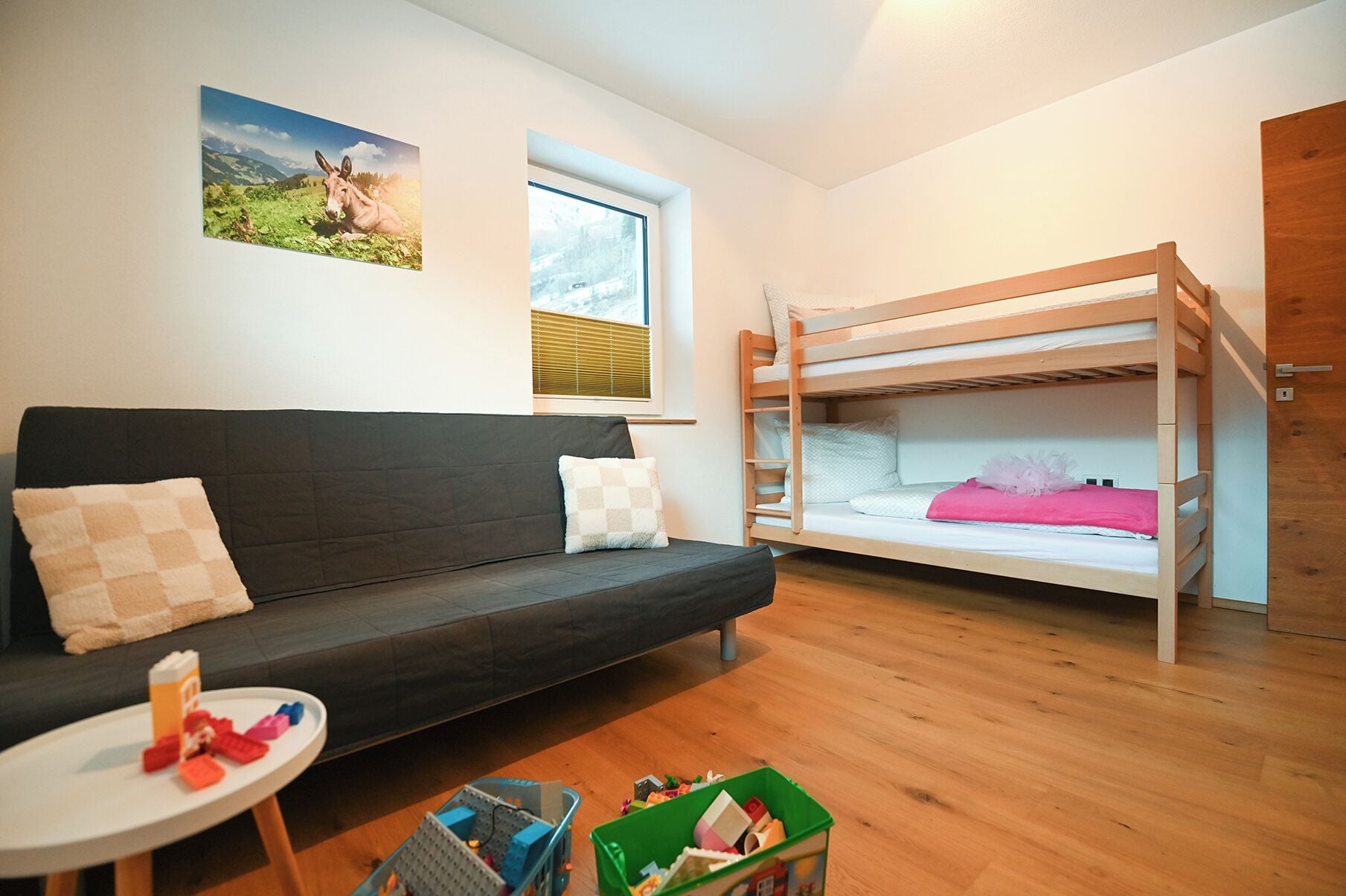 Ein Zimmer mit Etagenbett, einer Schlafcouch, einem kleinen Tisch mit Spielzeug und einem Fenster mit Jalousien.