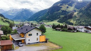 A mountain village with a prominent yellow building featuring a large 'G' logo, surrounded by lush green fields and mountains in the background.