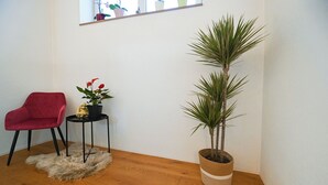 Et værelse med trægulv, en rød stol, et lille bord med en plante og en gylden figur, samt en høj plante i en vævet potte.