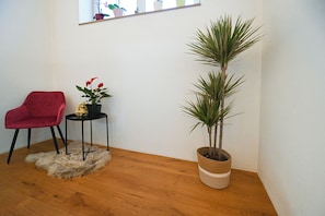 Ein Zimmer mit Holzboden, einem roten Sessel, einem kleinen Tisch mit Pflanze und goldener Figur sowie einer hohen Pflanze im gewebten Topf.