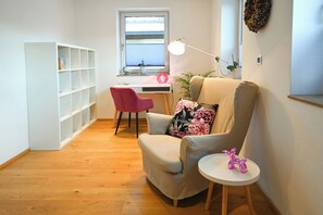 A room with a wooden floor, a beige armchair with a floral pillow, a pink chair, a white desk with a lamp, and a white shelving unit. - Chalet Creatina by we rent (Rauris)