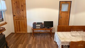 A room with a wooden floor, a desk with a computer and speakers, a table with a lace tablecloth, and wooden doors.