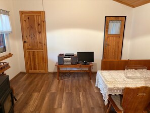 A room with a wooden floor, a desk with a computer and speakers, a table with a lace tablecloth, and wooden doors.