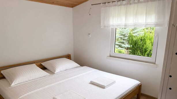 A double bed with white linens, a window with a view of greenery, and a blue rug on the floor.