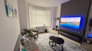 A living room with a sofa, a coffee table, a TV mounted on a wall, and a window with blinds. - Stylish 3BR House in Newham East London (London)