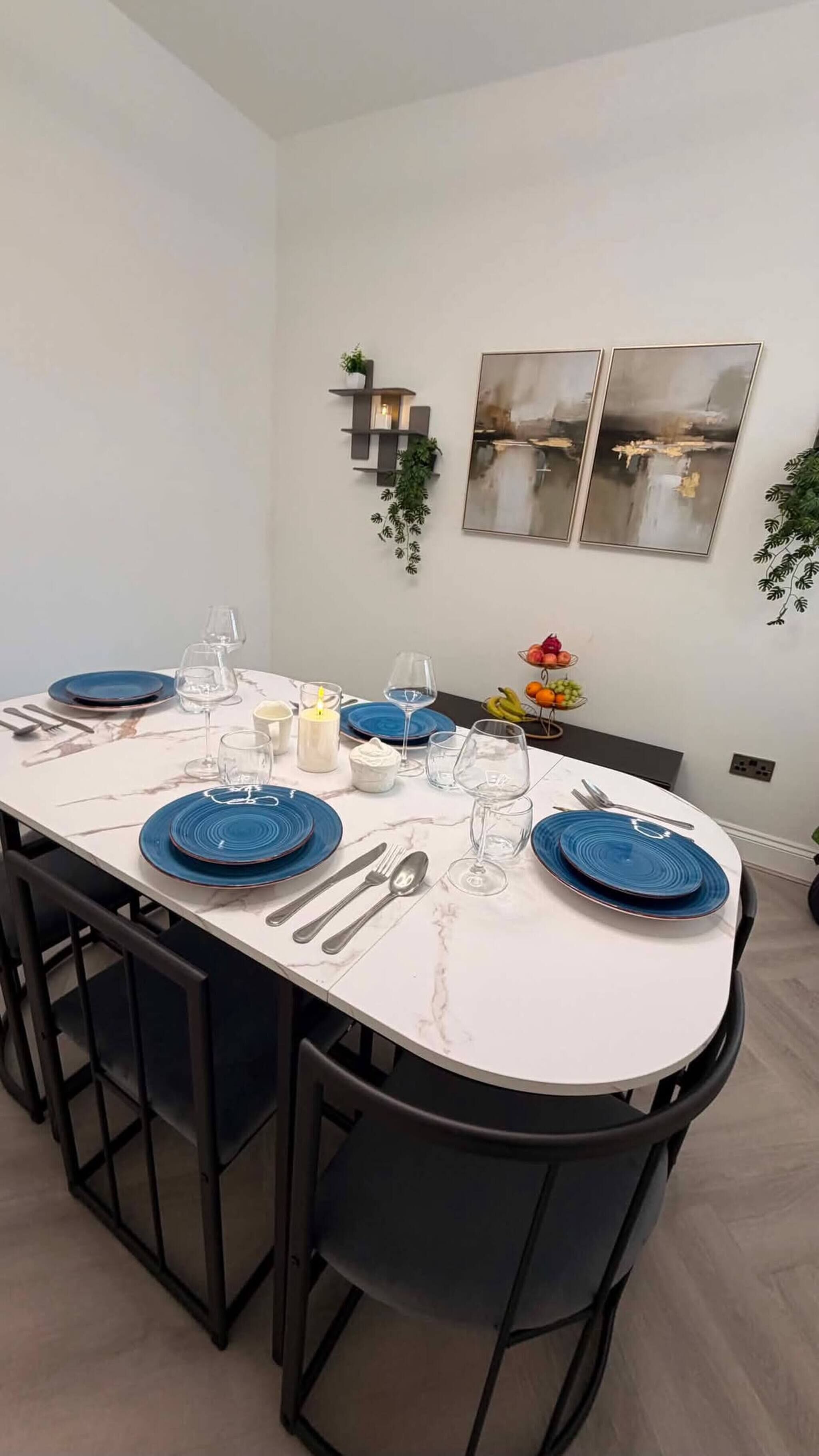 A dining area with a white marble table, blue plates, and black chairs. There are two wall-mounted shelves with plants and two framed pictures.