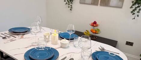 A dining area with a white marble table, blue plates, and black chairs. There are two wall-mounted shelves with plants and two framed pictures.