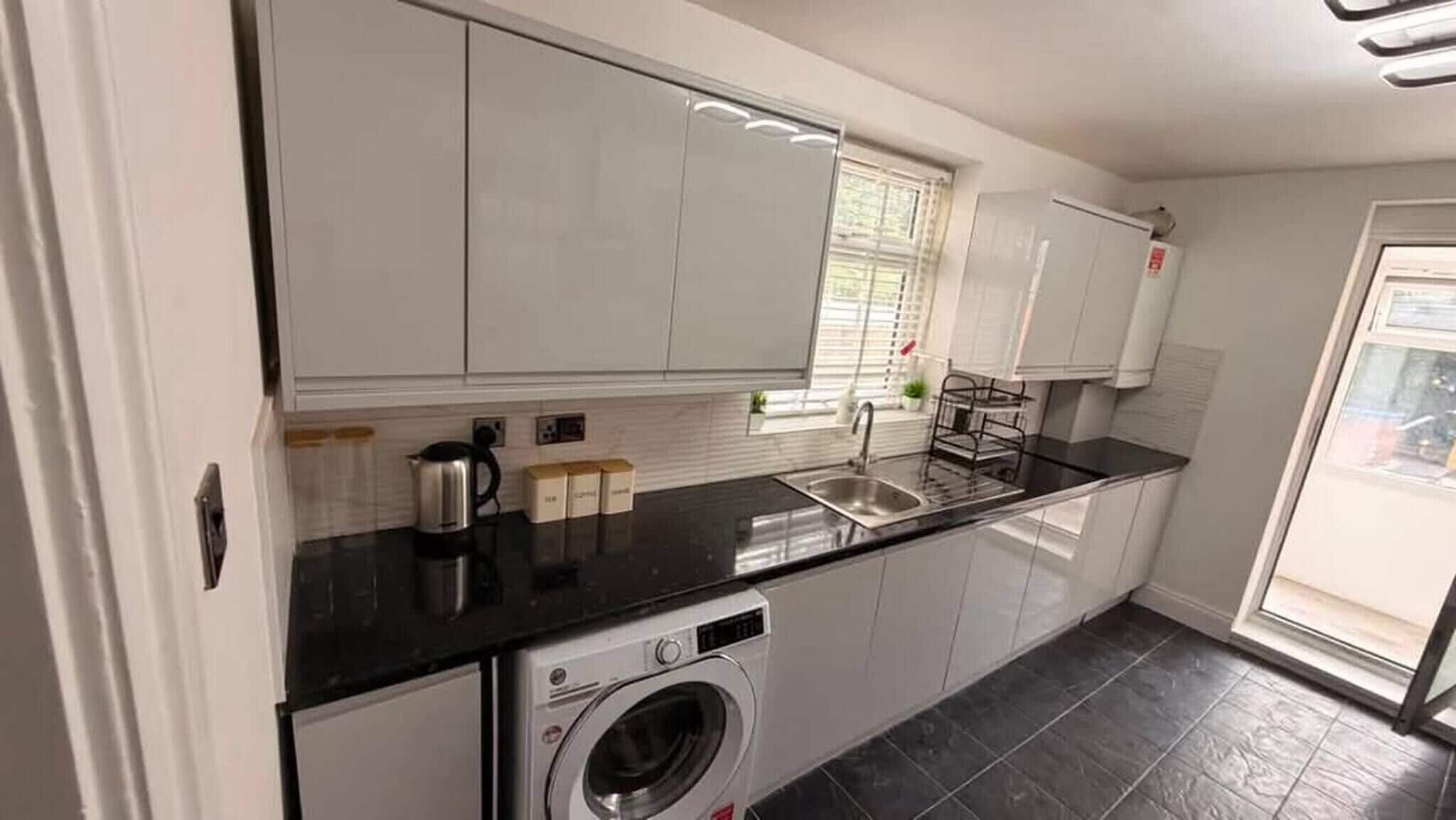 A modern kitchen with white cabinets, a black countertop, a built-in washing machine, and a window with blinds.