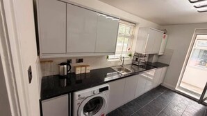 A modern kitchen with white cabinets, a black countertop, a built-in washing machine, and a window with blinds. - Stylish 3BR House in Newham East London (London)