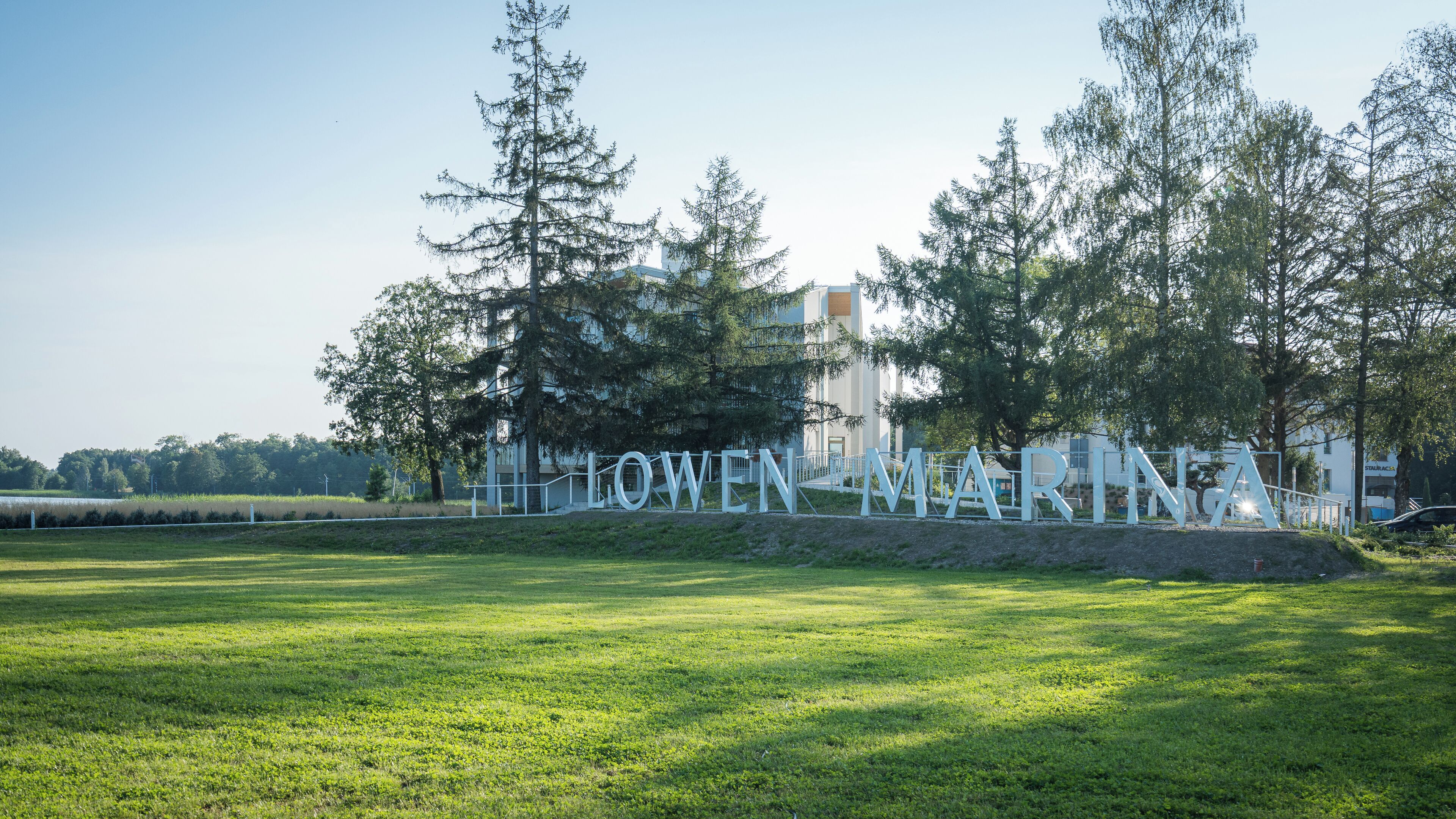 A sign with the text 'LOWEN MARINA' in a grassy field with trees and a building in the background.