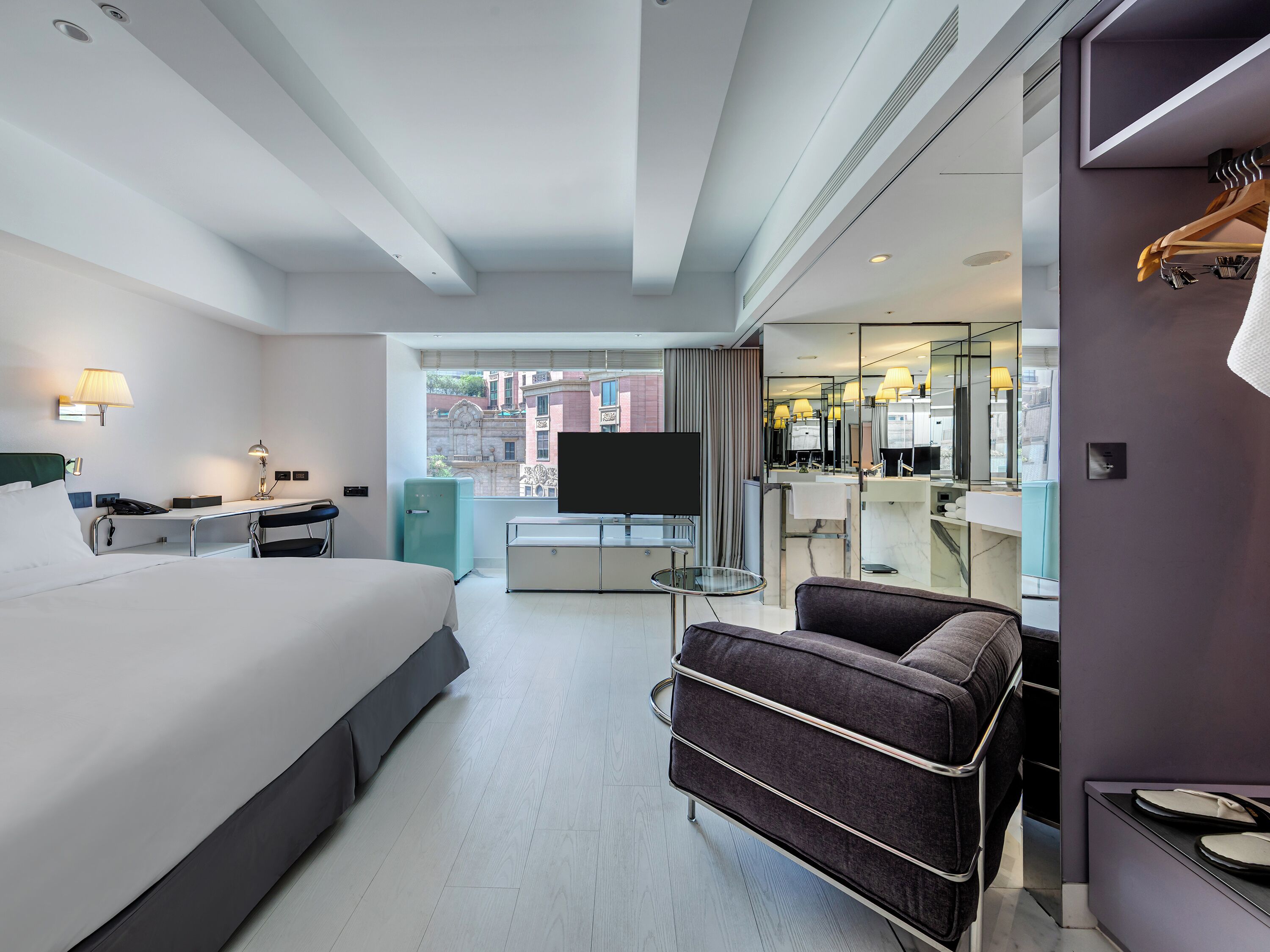 A modern hotel room with a bed, desk, chair, TV, and a view of the city.