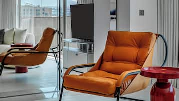 A modern hotel room with a large window, a sofa, two orange armchairs, a flat-screen TV, and a red side table.