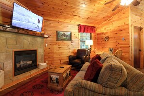 Ein gemütliches Blockhauss-Interieur mit Kamin aus Stein, einem an der Wand befestigten Fernseher, einem Ledersessel, einer Couch und einer Holzbalkendecke.