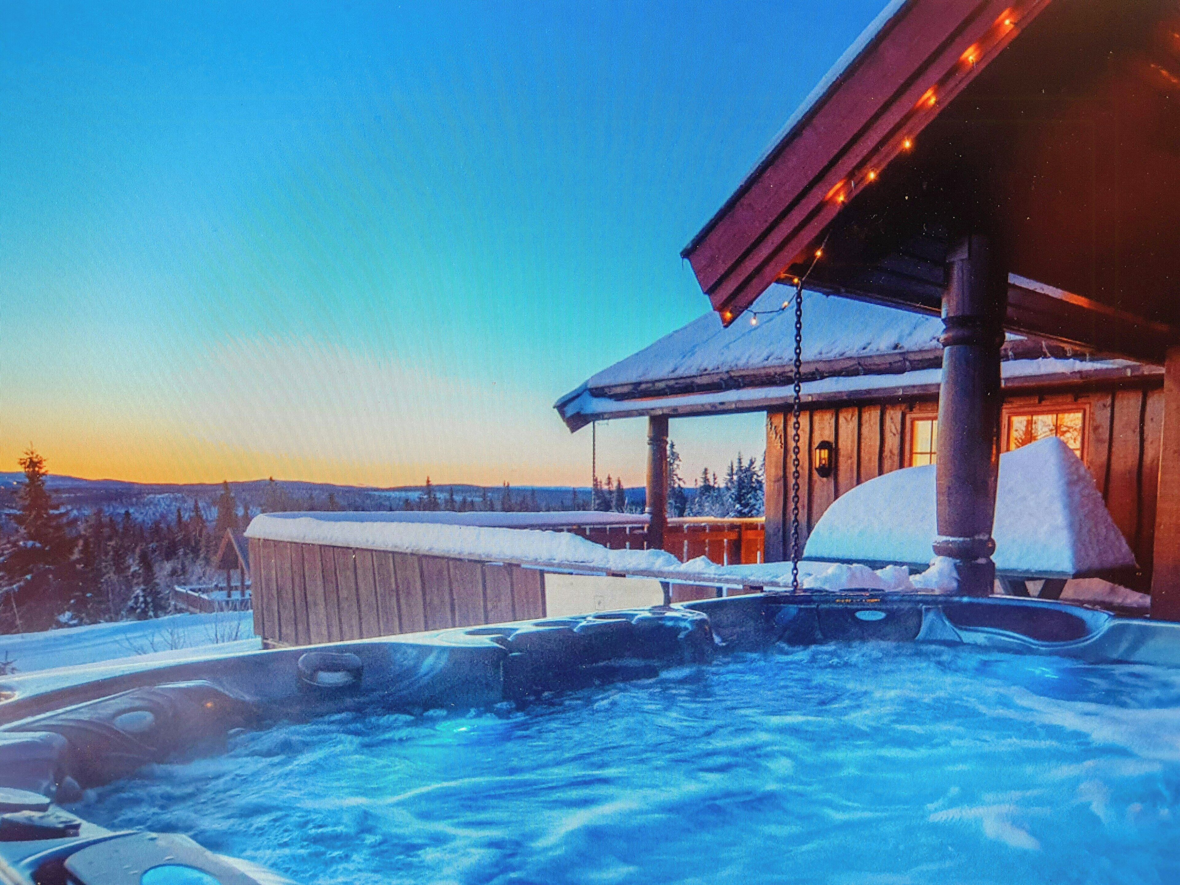 A hot tub with a wooden deck, a wooden cabin with a snow-covered roof, and a snowy landscape in the background.