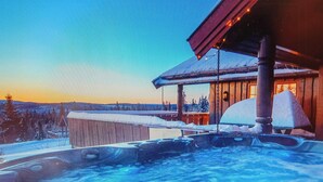 Un jacuzzi con terraza de madera, una cabaña de madera con tejado cubierto de nieve y un paisaje nevado al fondo.
