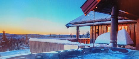 A hot tub with a wooden deck, a wooden cabin with a snow-covered roof, and a snowy landscape in the background.
