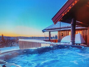 Ein Whirlpool mit Holzterrasse, eine Holzhütte mit schneebedecktem Dach und verschneite Landschaft im Hintergrund.